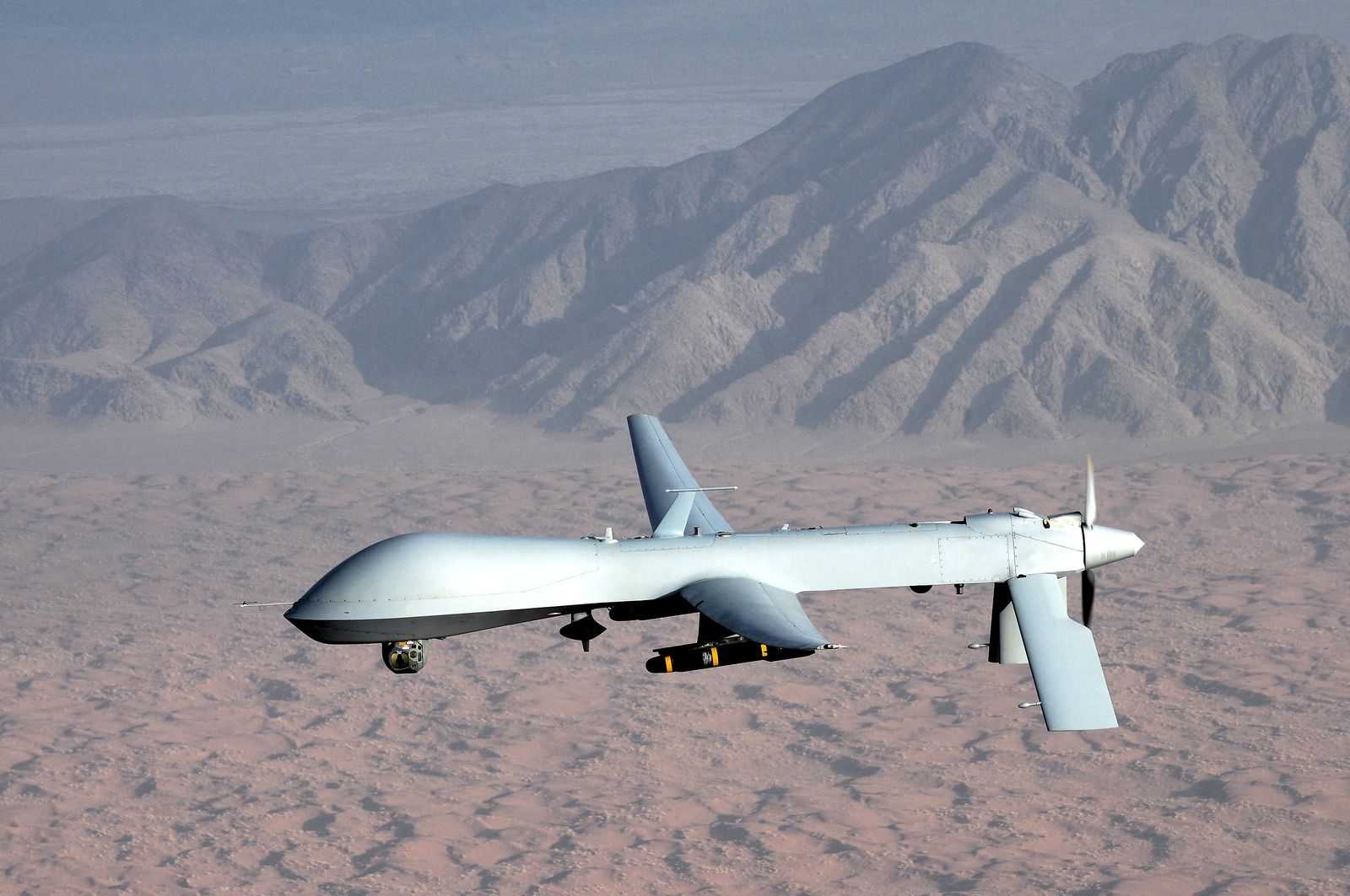 Predator drone photographed from an aircraft flying alongside. The landscape that can be seen could be a desert. In the background, a small mountain range can be seen. 