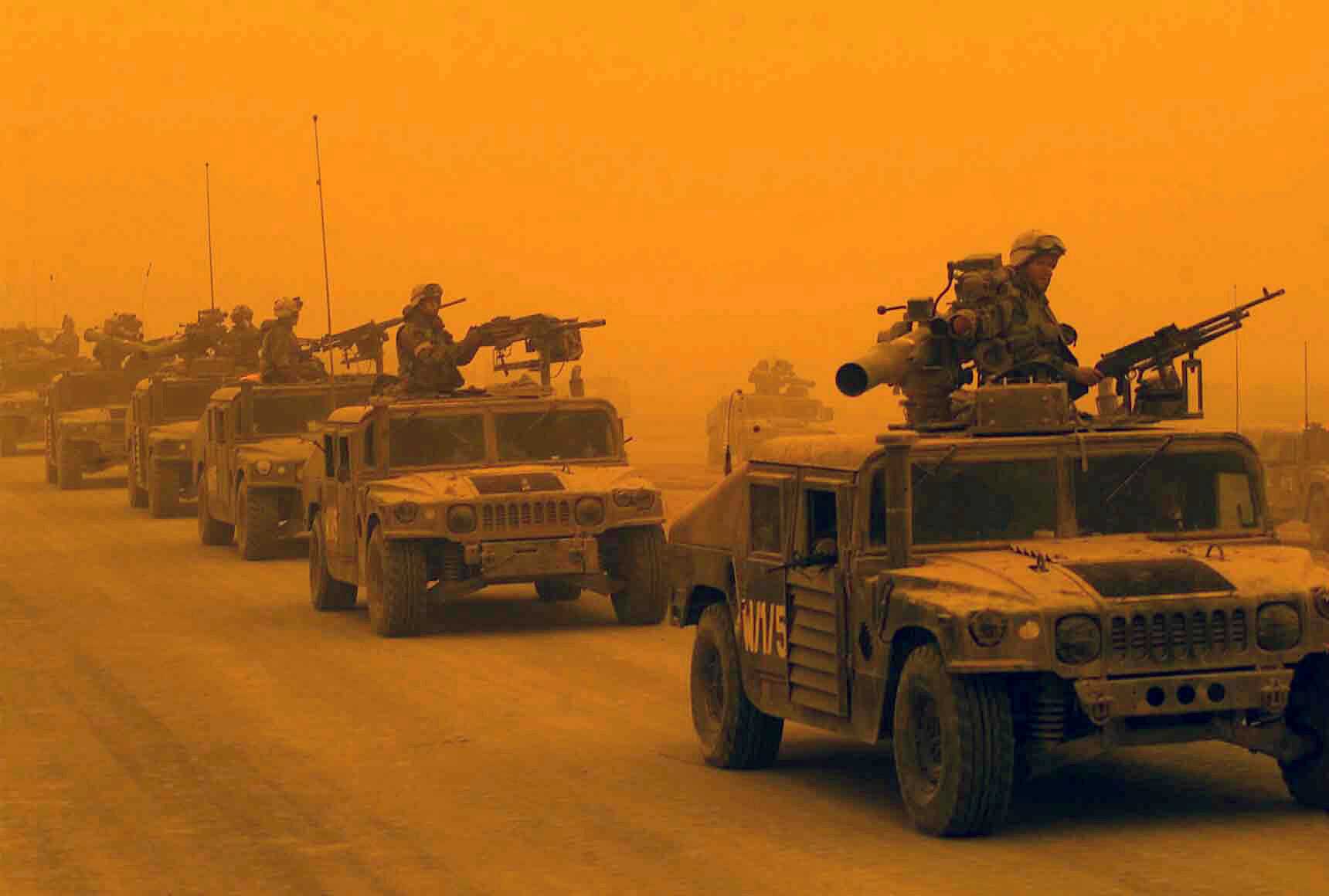 The image, taken in 2003 during the Iraq War, shows a convoy of military Humvees driving through a desert landscape under an intense orange haze, likely caused by a sandstorm. Soldiers in full combat gear are positioned on top of the vehicles, manning large mounted machine guns and other weaponry. The lead vehicle has a visible marking 'KMV5' on its side. The convoy appears heavily armed and moving in formation on a dusty road, emphasizing a tense and hostile environment. 