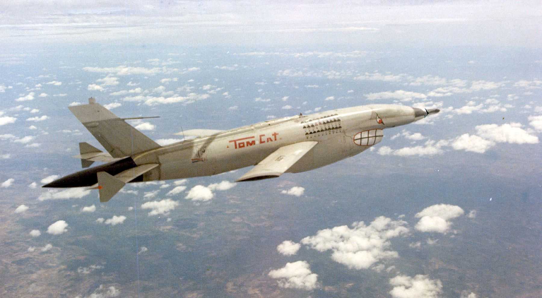 drone flies high above the ground over some clouds. The drone resembles a fighter plane. It is labelled ‘Tom Cat’ and has a grim face painted on it. The ground could be a forest. 