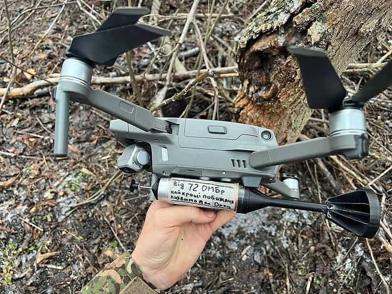 A hand holding a quadcopter drone with a granade attached to it. The main body of the drone is slightly larger than the hand holding it