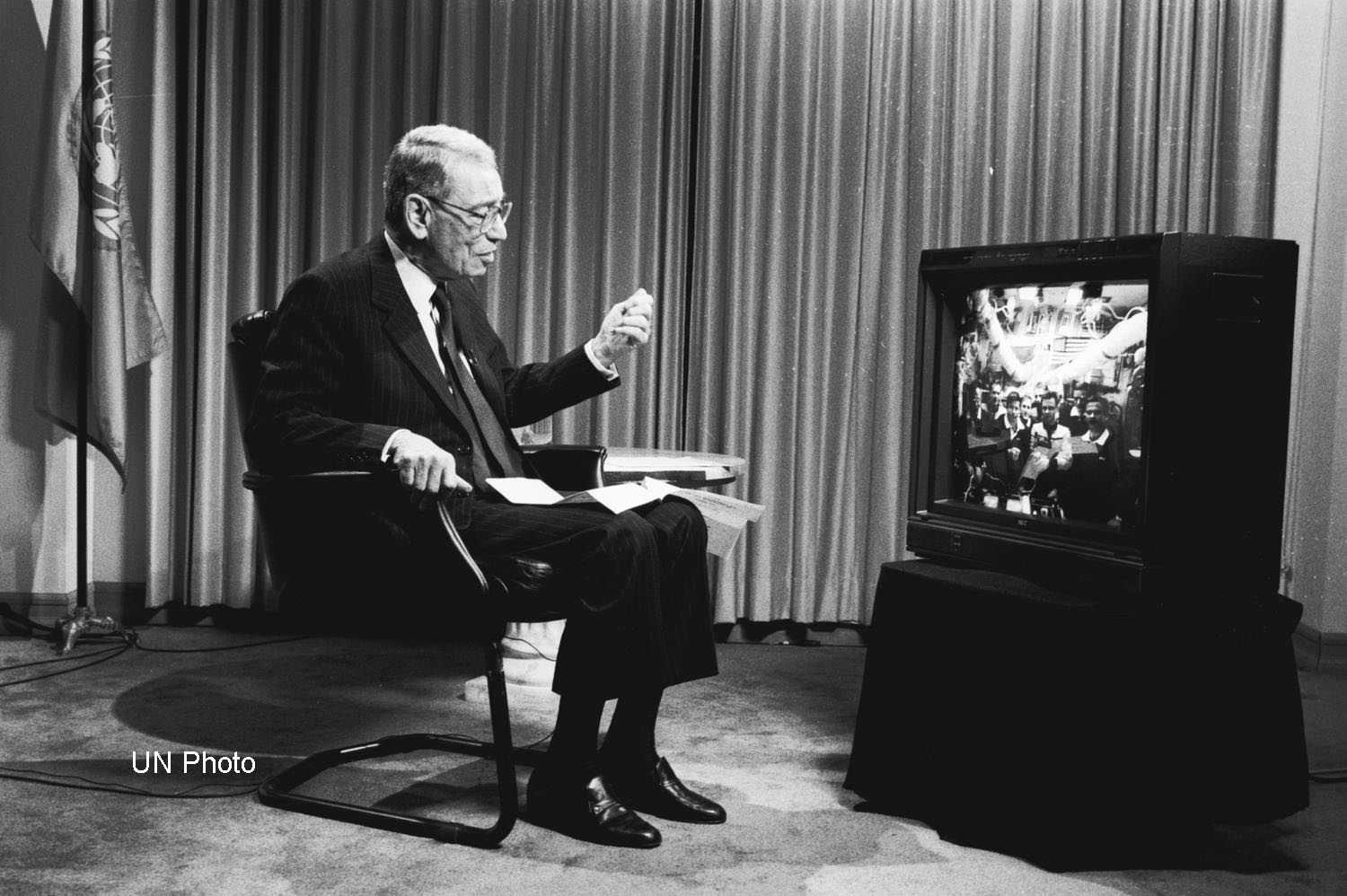 Black and white photo of Secretary-General Boutros Boutros-Ghali sitting in front of a TV with the UN flag in the background, celebrating the 50th anniversary of the UN with the Shuttle-Mir docking mission on  November 17, 1995