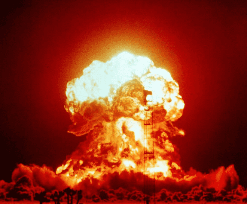 A massive nuclear explosion dominates the scene, captured at the moment of detonation. The image shows an enormous, fiery mushroom cloud rising into the dark sky, radiating intense red, orange, and yellow hues. The base of the explosion is surrounded by billowing, glowing clouds of smoke and fire. Silhouetted in front of the blast are small trees and a tall, thin tower structure, giving a sense of scale to the powerful eruption. The brightness at the core of the blast contrasts starkly with the dark surroundings, emphasizing the energy and intensity of the explosion.